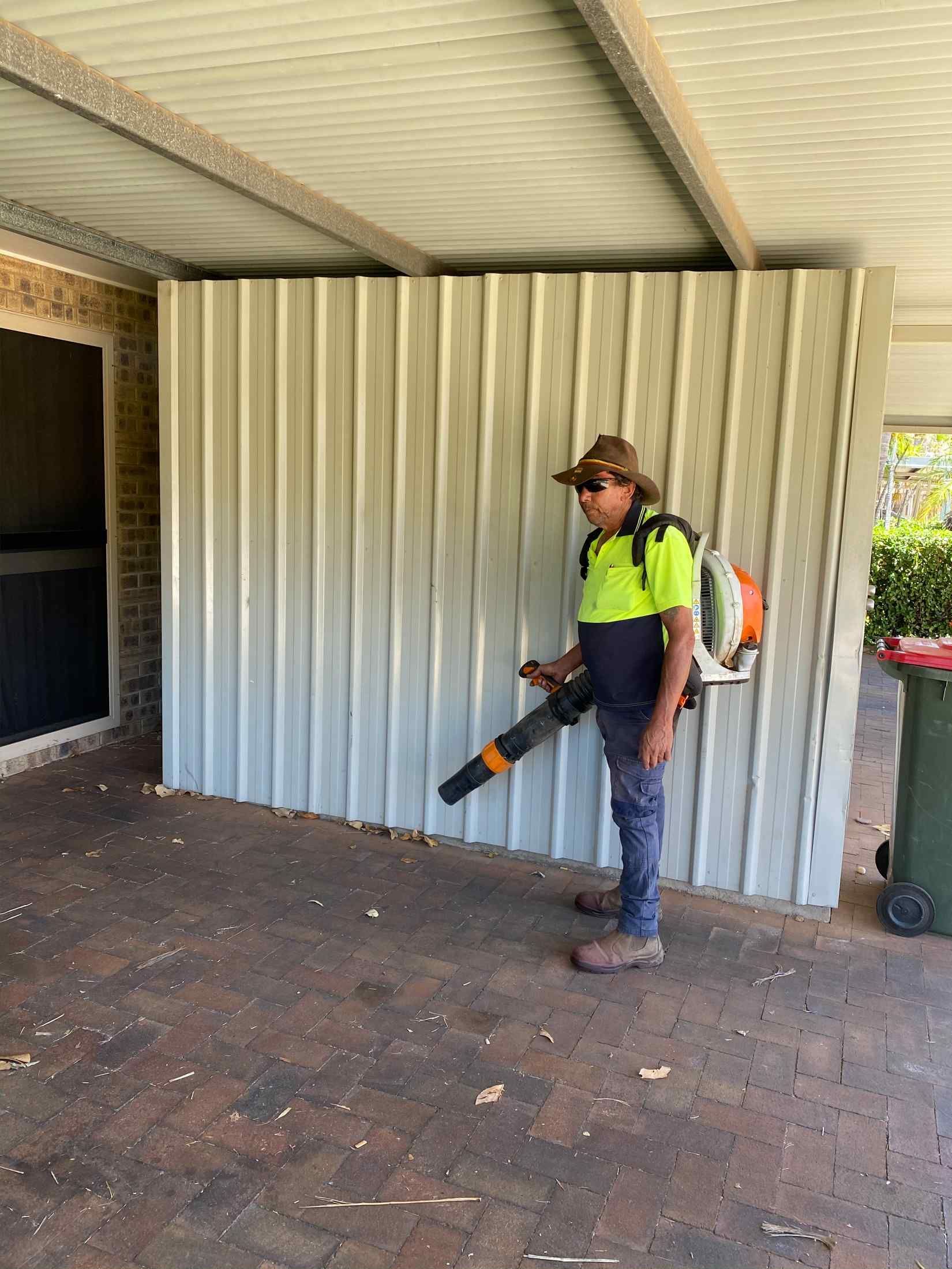 Person Using a Leaf Blower on a Concrete Patio Next to a Metal Wall and Trash Can — Darrell's Mowing Services in Sunset, QLD