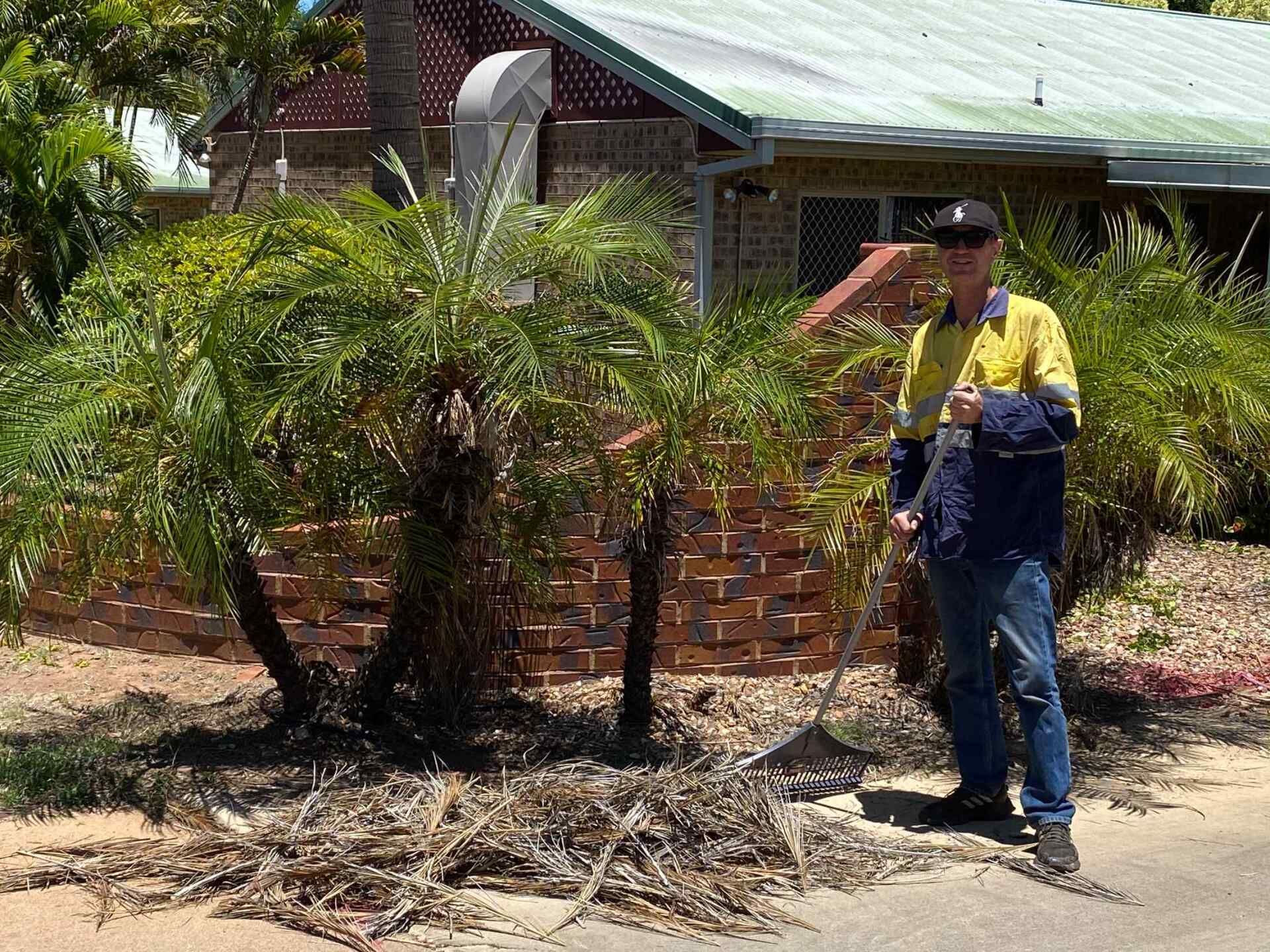 Man Raking Palm Tree Debris in Front of a Brick House — Darrell's Mowing Services in Sunset, QLD