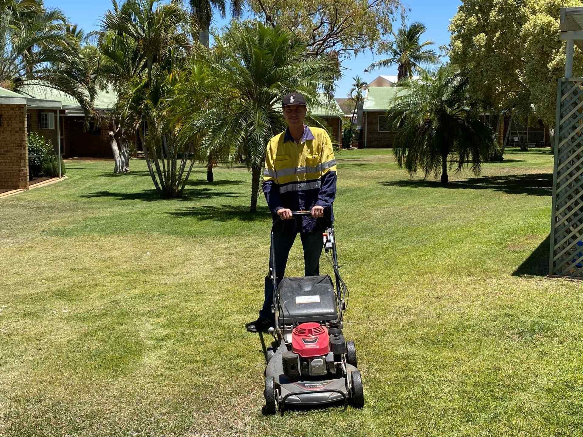 Man in Workwear Mowing a Grassy Lawn With a Red and Black Lawnmower — Darrell's Mowing Services in Sunset, QLD