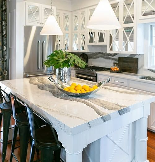 Bright white kitchen island with marble top, pendant lights, stainless fridge, and fruit bowl centerpiece