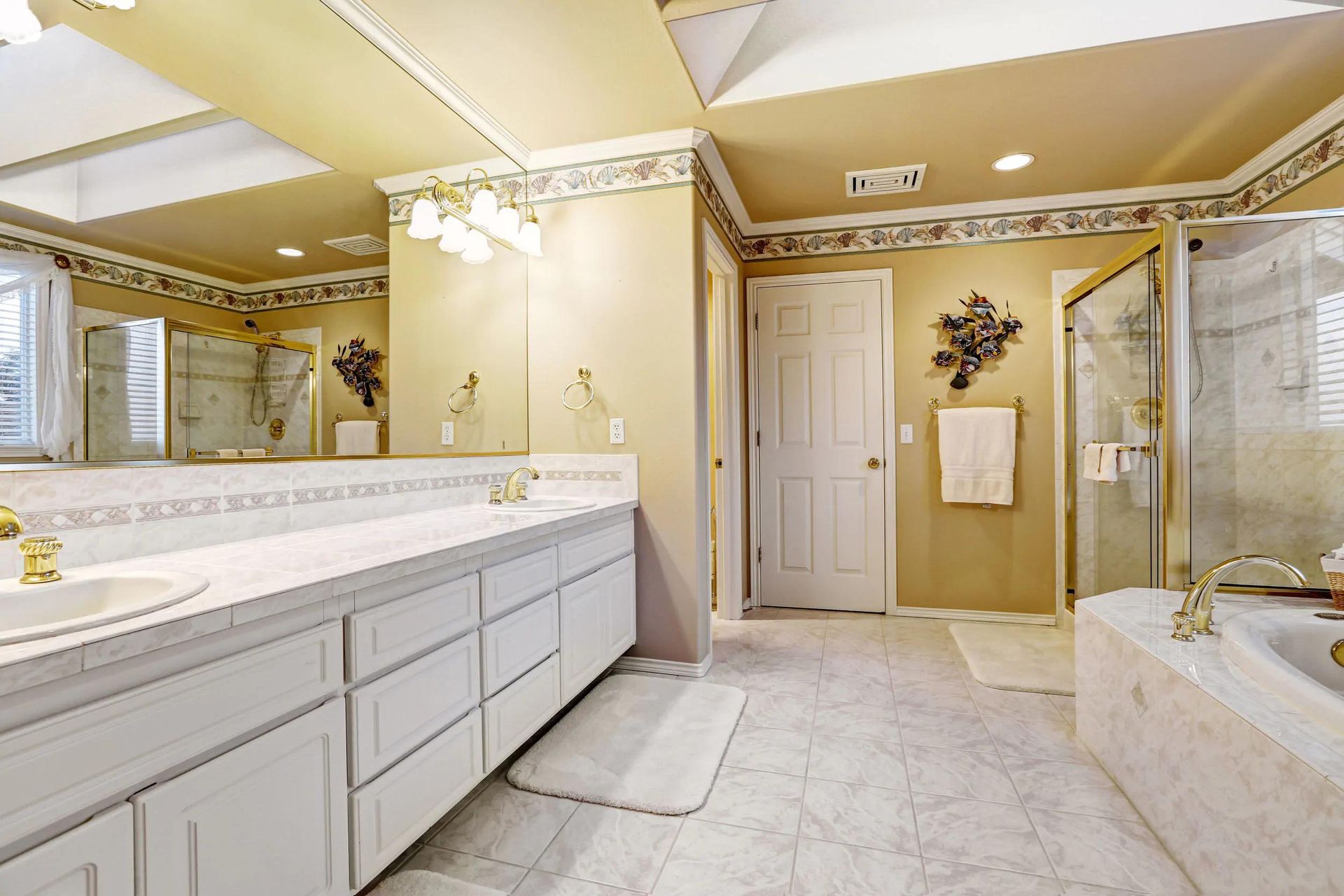 Bright beige bathroom with double vanity, large mirror, chandelier, and tiled tub area