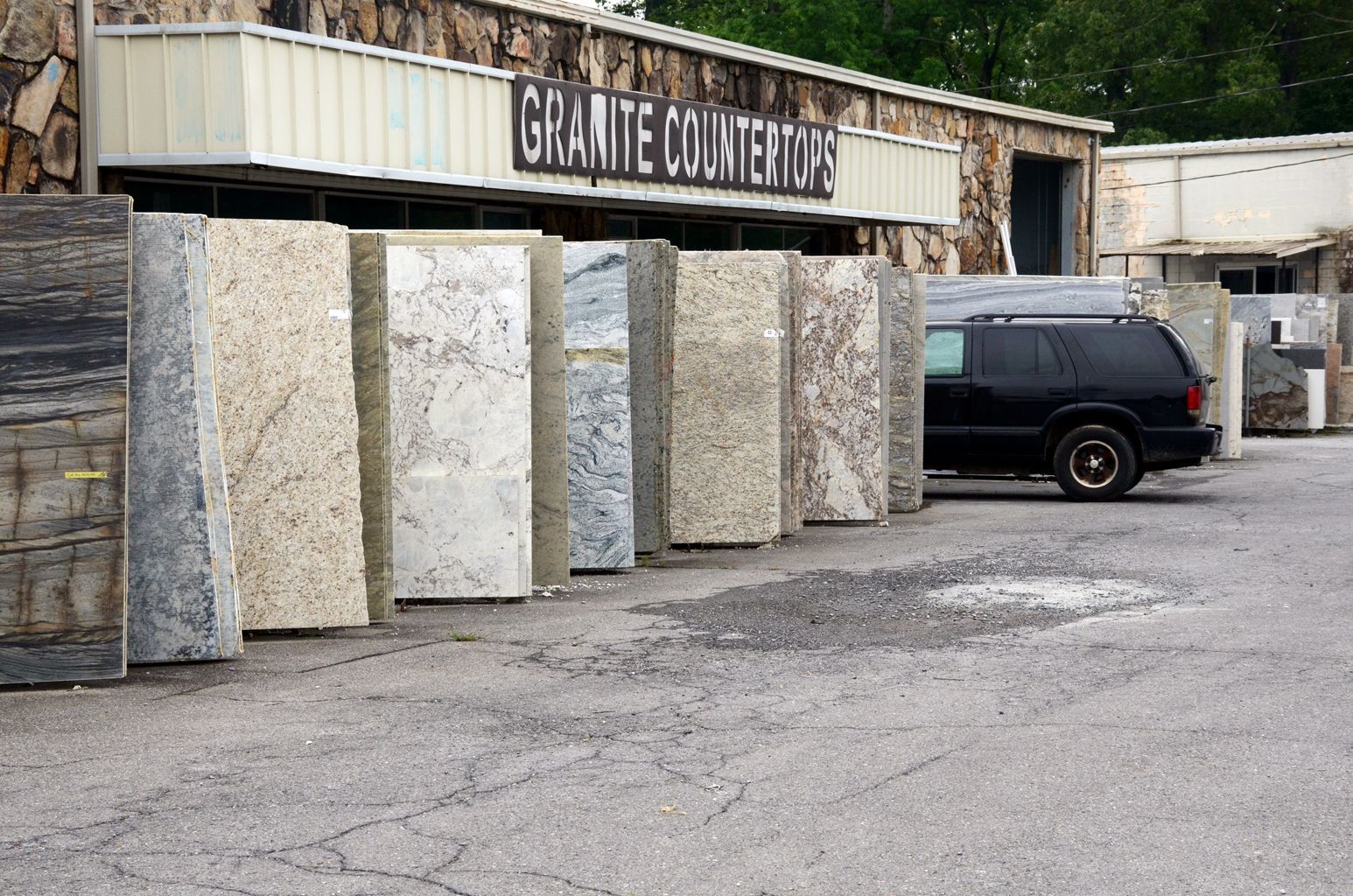Granite countertop yard with large stone slabs displayed outside a warehouse and a black van parked nearby