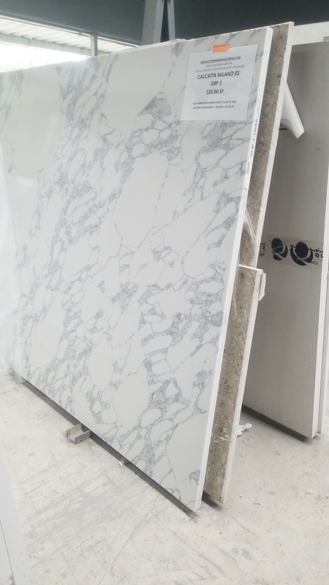Large white marble-look slab leaning upright in a warehouse or showroom, with a small label at the top right.
