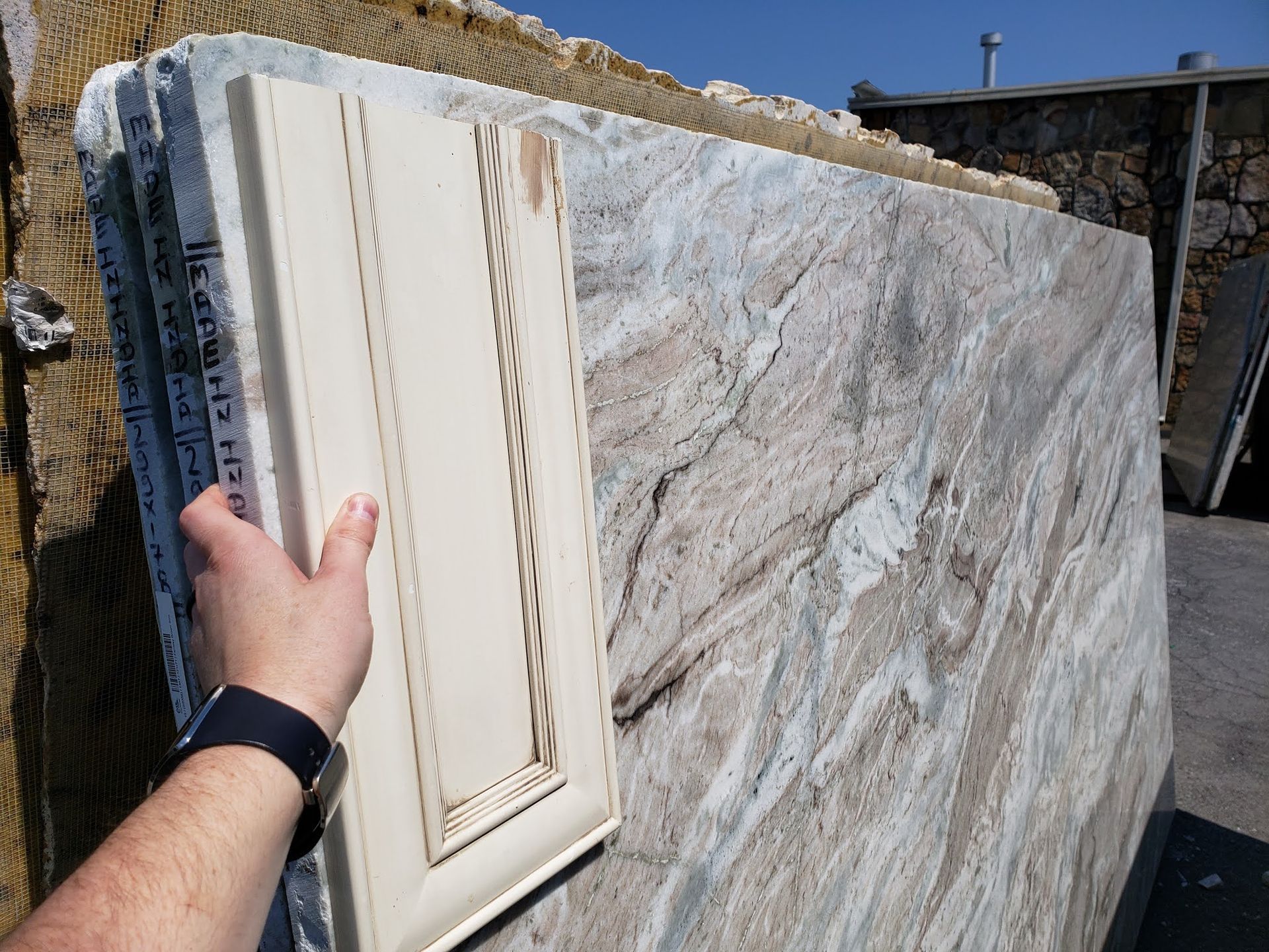 Hand holding a white cabinet door against a large polished stone slab outdoors