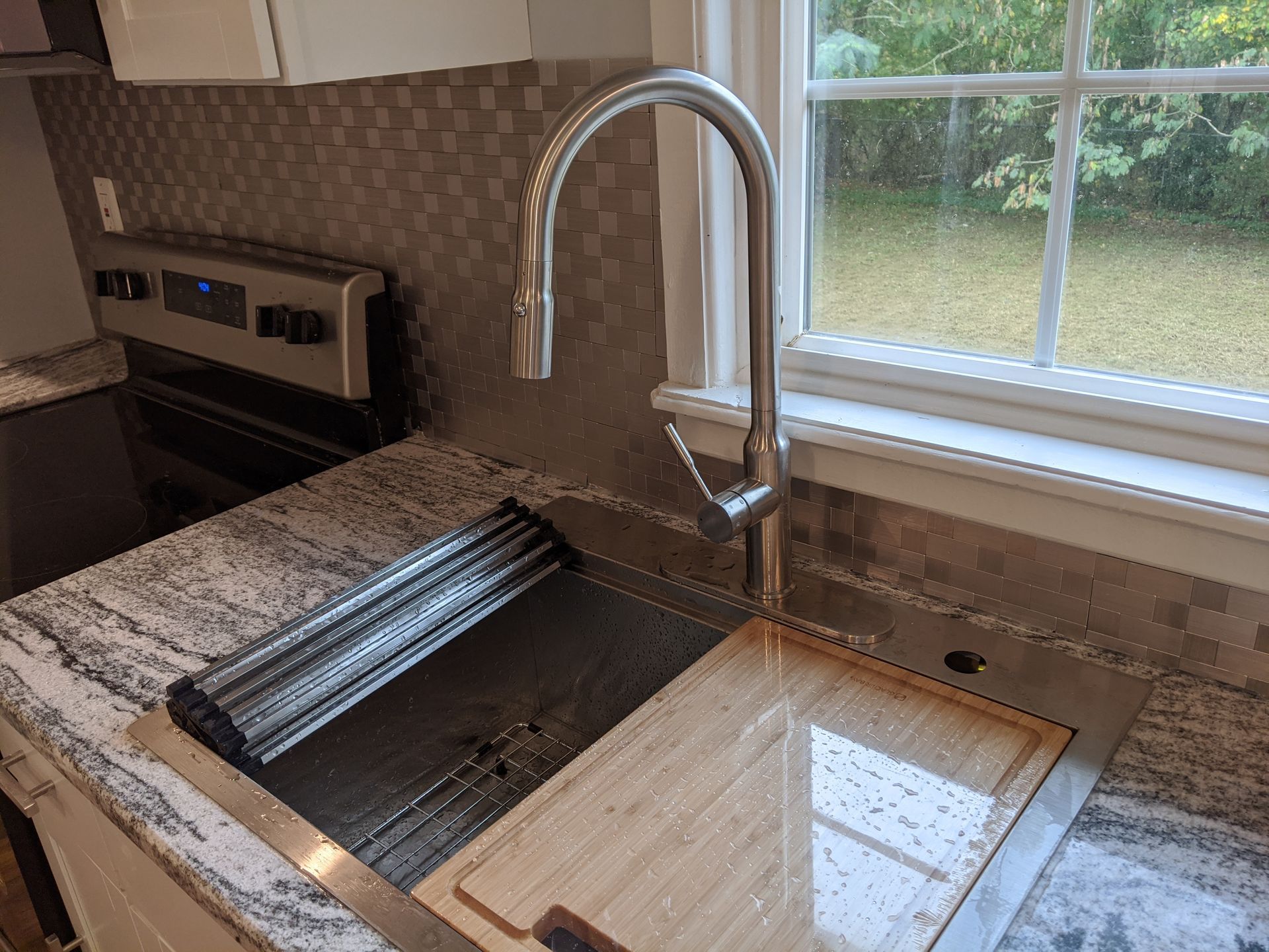 Modern kitchen sink with chrome faucet, granite countertop, and window view of greenery