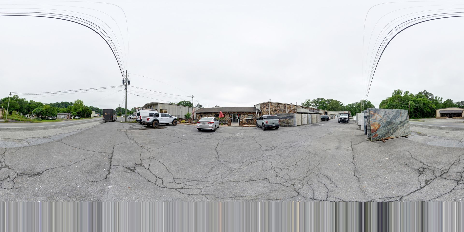 Wide gravel parking lot with several parked vehicles, low buildings, and trees under an overcast sky.