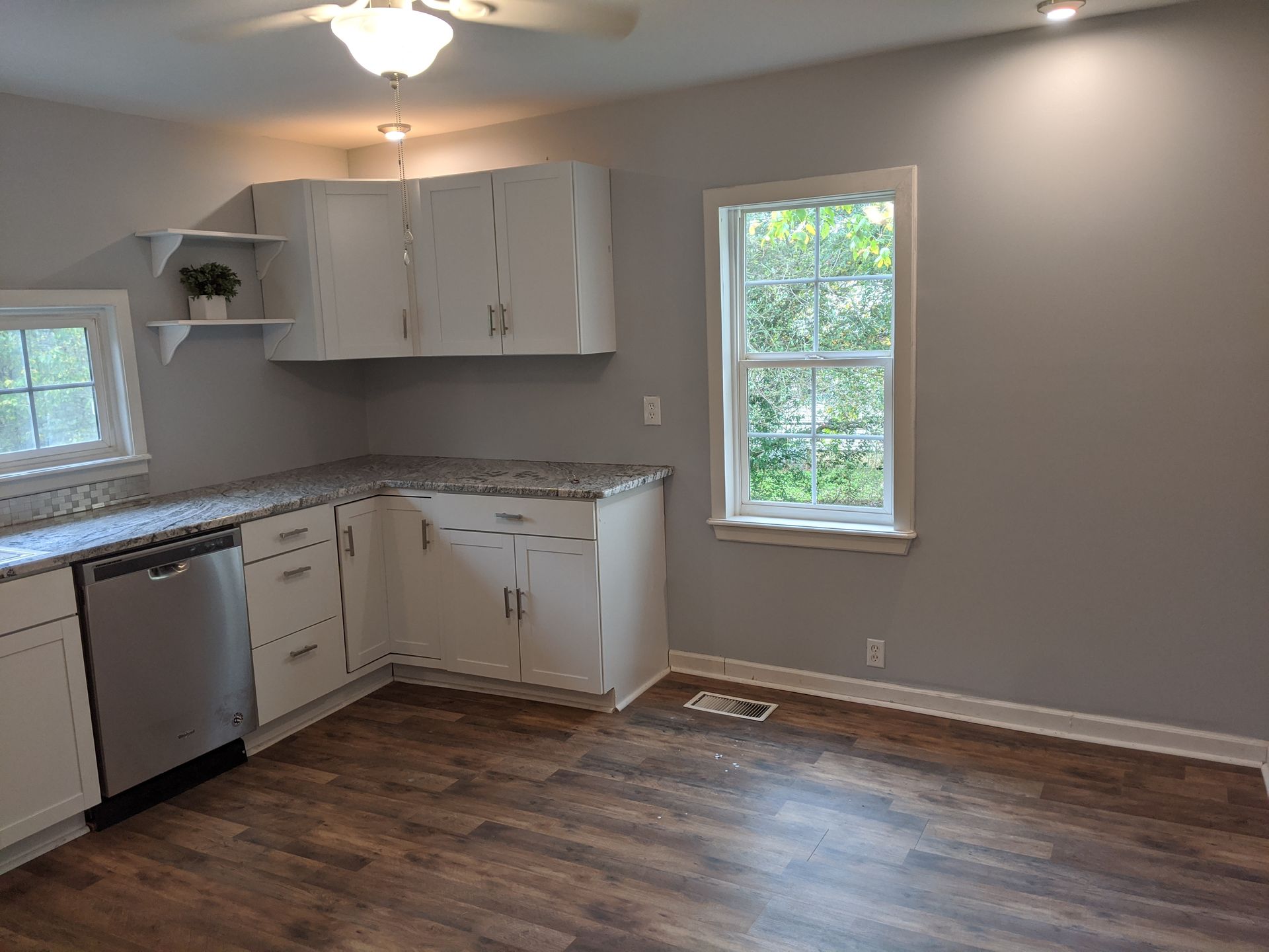Empty kitchen with white cabinets, stainless dishwasher, wood floors, and a window over the baseboard heater.