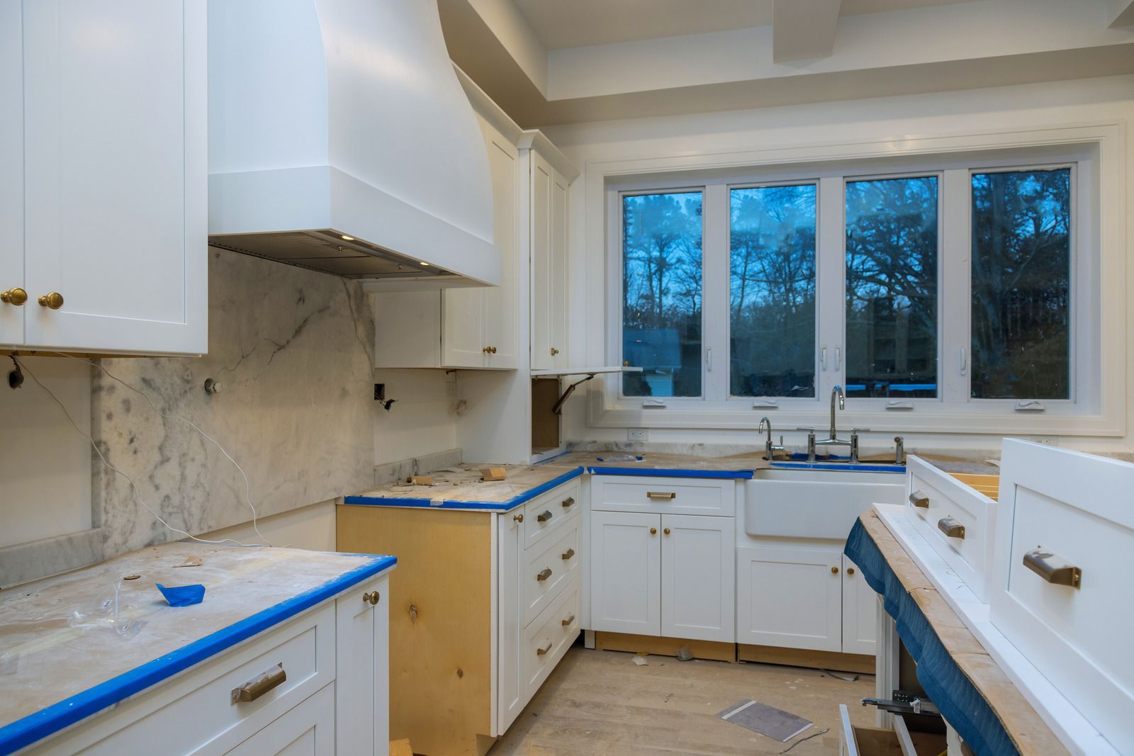 Bright kitchen under renovation with white cabinets, blue-taped countertops, and a large window above the sink