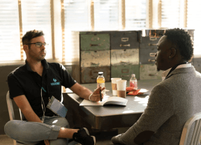 Two people talking at a table in a bright café, with drinks and a laptop open.
