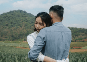 Two people embracing in a grassy field with a green hill in the background.