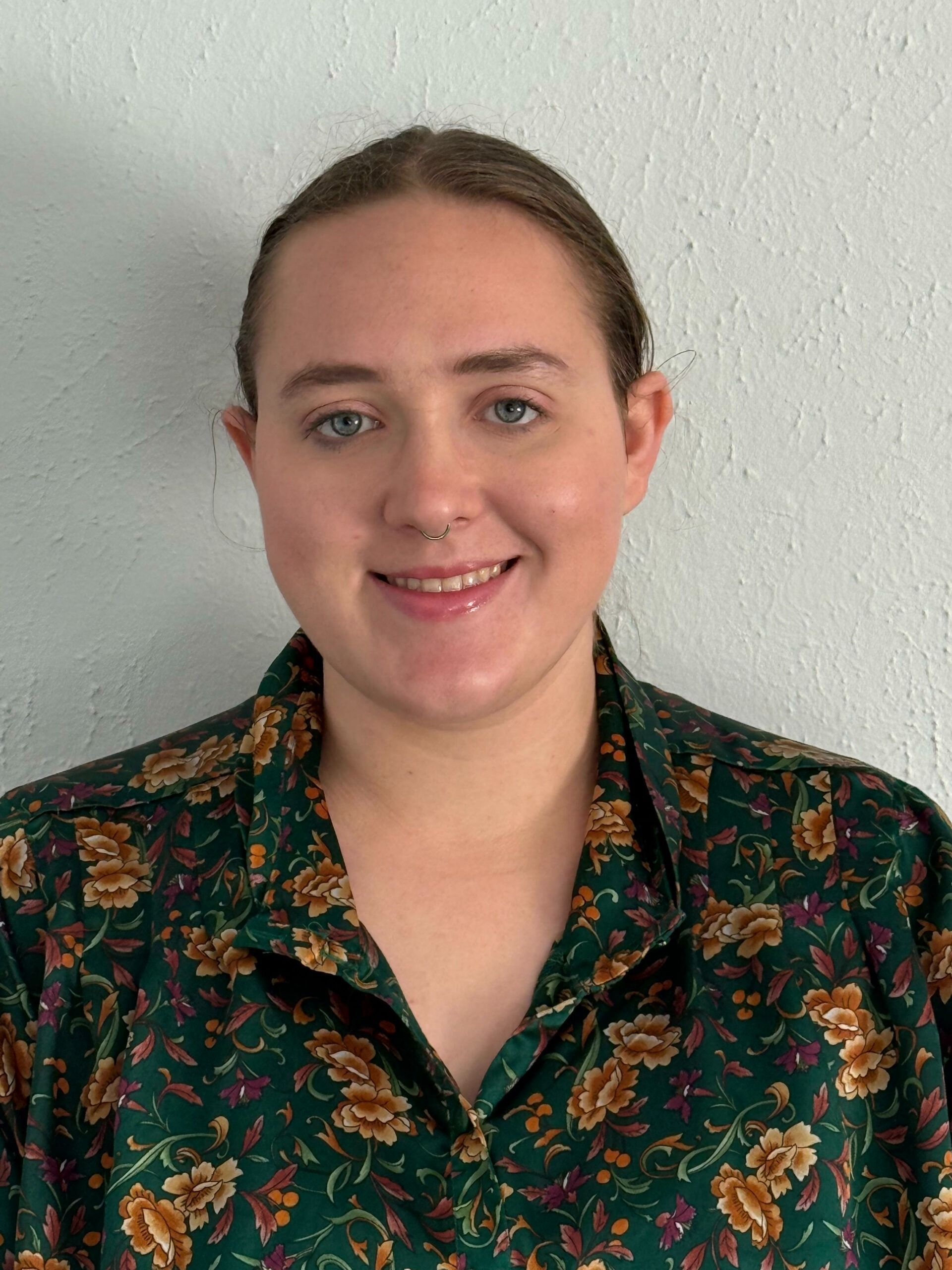 Smiling person in a dark floral shirt standing against a light gray wall