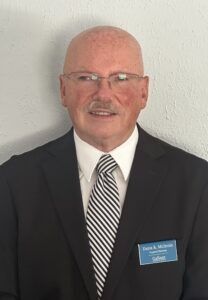 Man in a black suit, white shirt, and striped tie wearing a name badge, standing against a white wall.