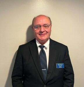 Man in a dark suit, tie, and glasses standing against a plain wall with a name badge.
