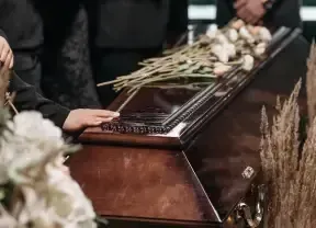 Hands resting on a polished coffin adorned with white flowers and beige stalks during a funeral.