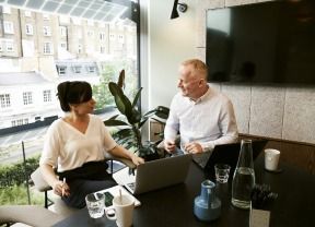 Two people chatting at a table with a laptop in a bright office lounge area.