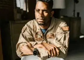 Man in tan military uniform leaning on a table with folded arms, looking ahead in a dim room