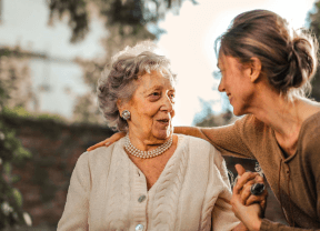 Two women smiling and talking outdoors, one older and one younger, with trees blurred in the background