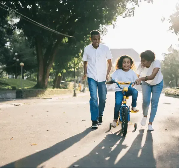 Three people walking with a child riding a blue bicycle on a sunlit park path