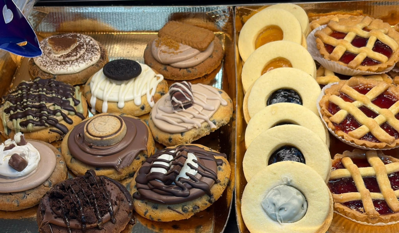 Vassoio di ciambelle e pasticcini assortiti decorati, tra cui ciambelle ad anello e torte a griglia, in una vetrina da pasticceria.