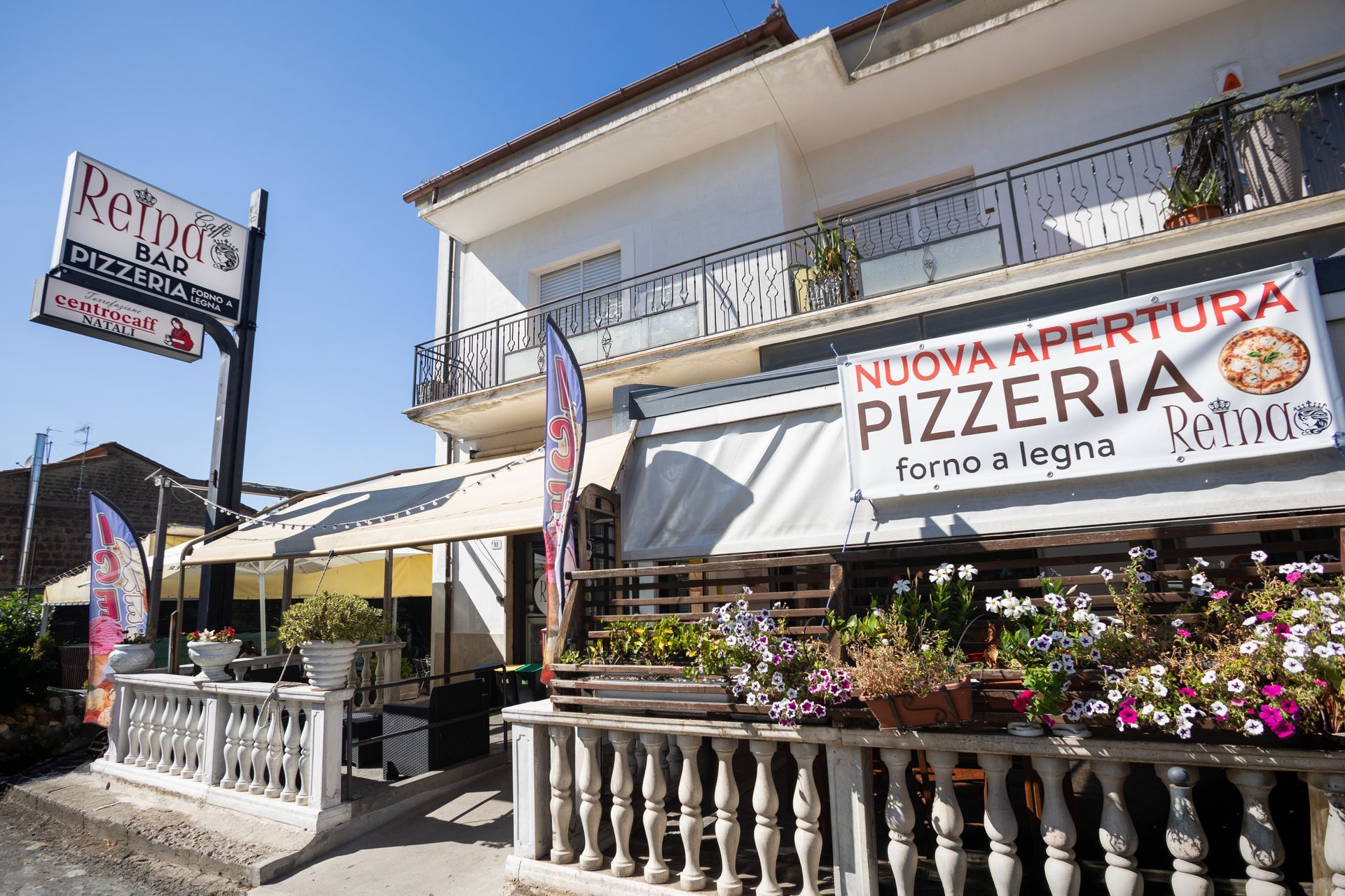 Pizzeria sul ciglio della strada con tende da sole bianche, balcone e fiori colorati sotto un cielo azzurro e soleggiato.