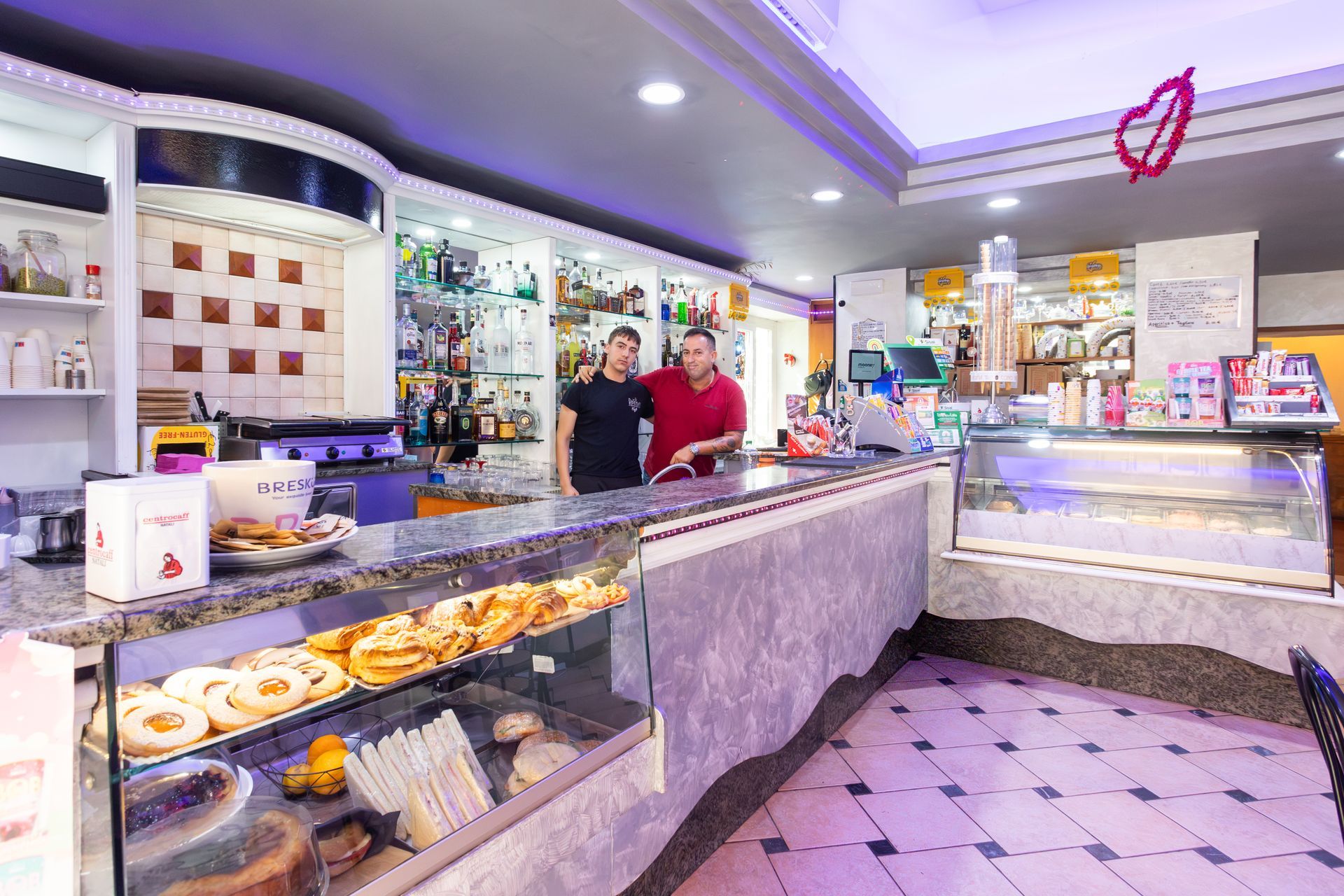 Bancone luminoso della pasticceria con pasticcini, vetrine espositive e due addetti dietro il bancone