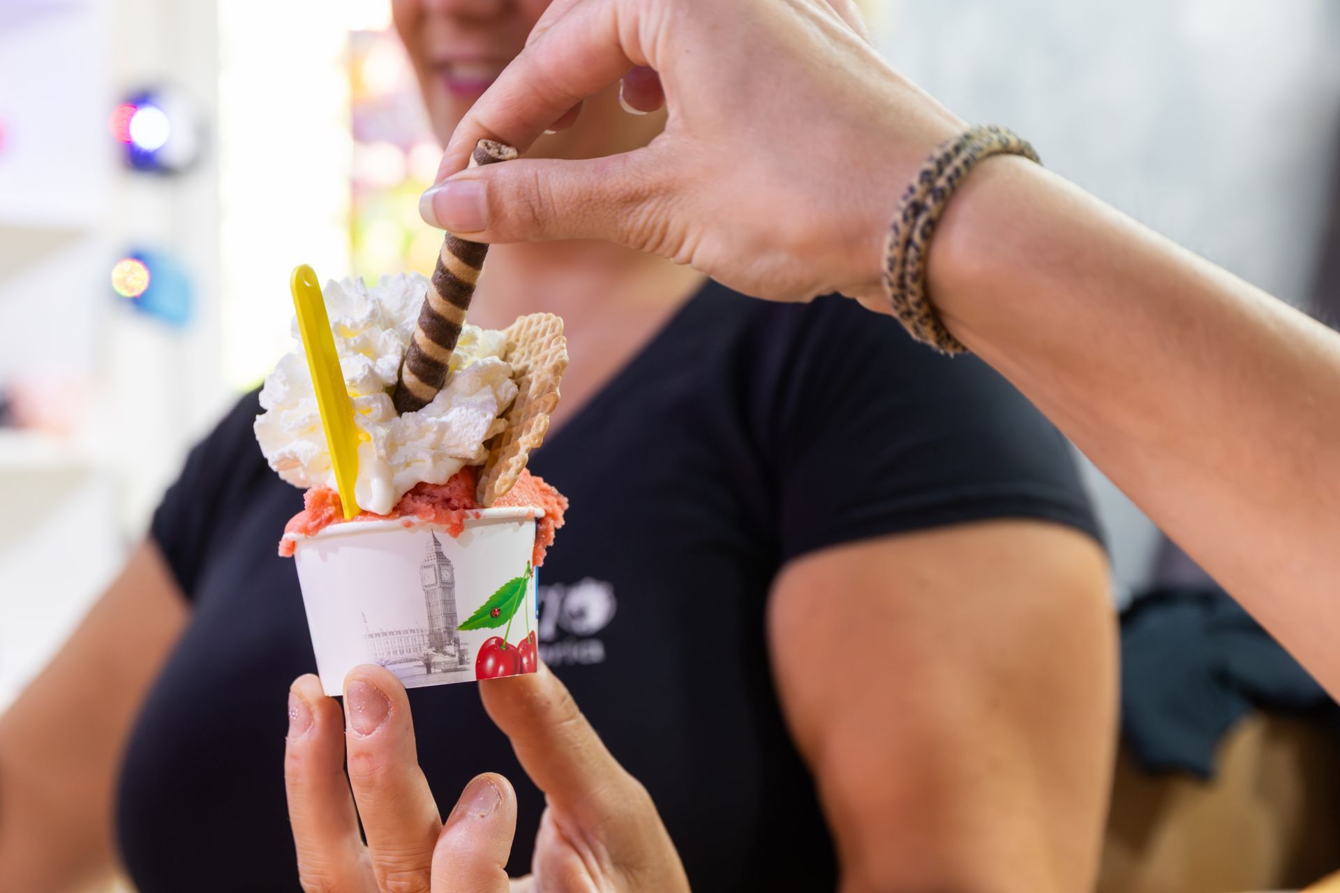 Mani che reggono una colorata coppetta da dessert con panna montata e bastoncini di wafer durante un evento all'aperto.