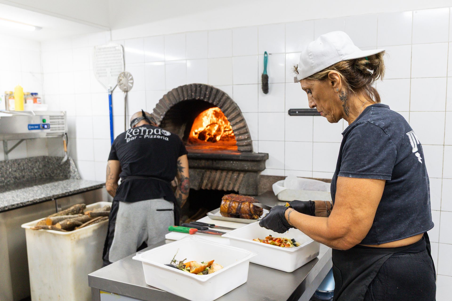 Due fornai impiattano il cibo accanto a un forno a legna in una cucina piastrellata.