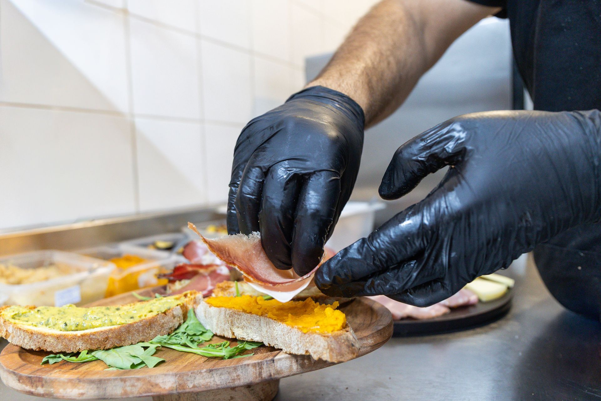 Mani guantate che assemblano un panino con lattuga, formaggio e prosciutto su un bancone della cucina.