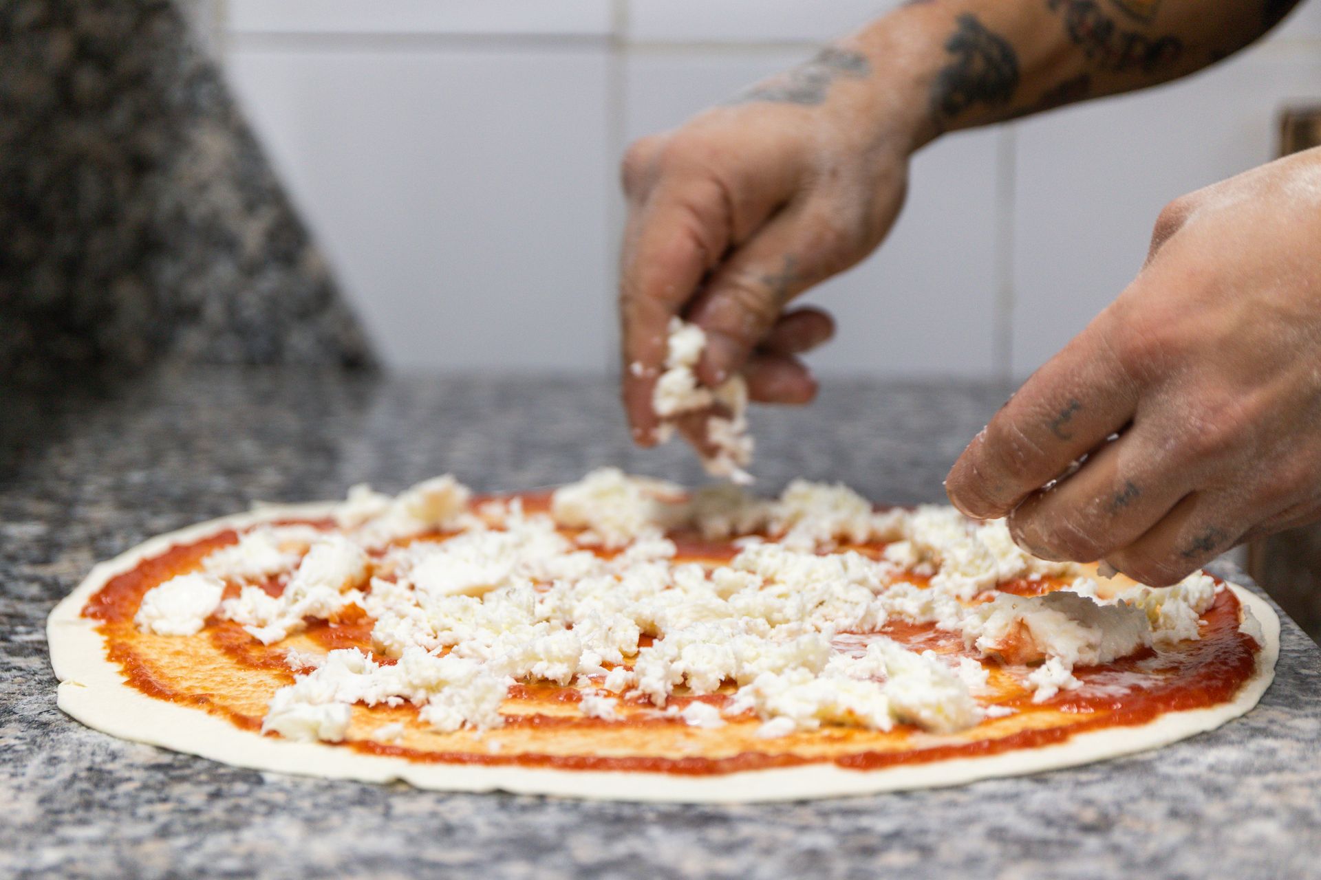 Delle mani cospargono di formaggio una base di pizza cruda su un piano di lavoro.