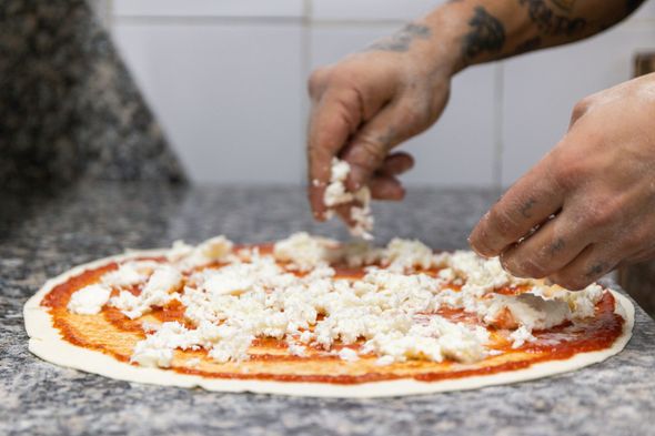 Delle mani cospargono di formaggio una base di pizza cruda su un piano di lavoro.