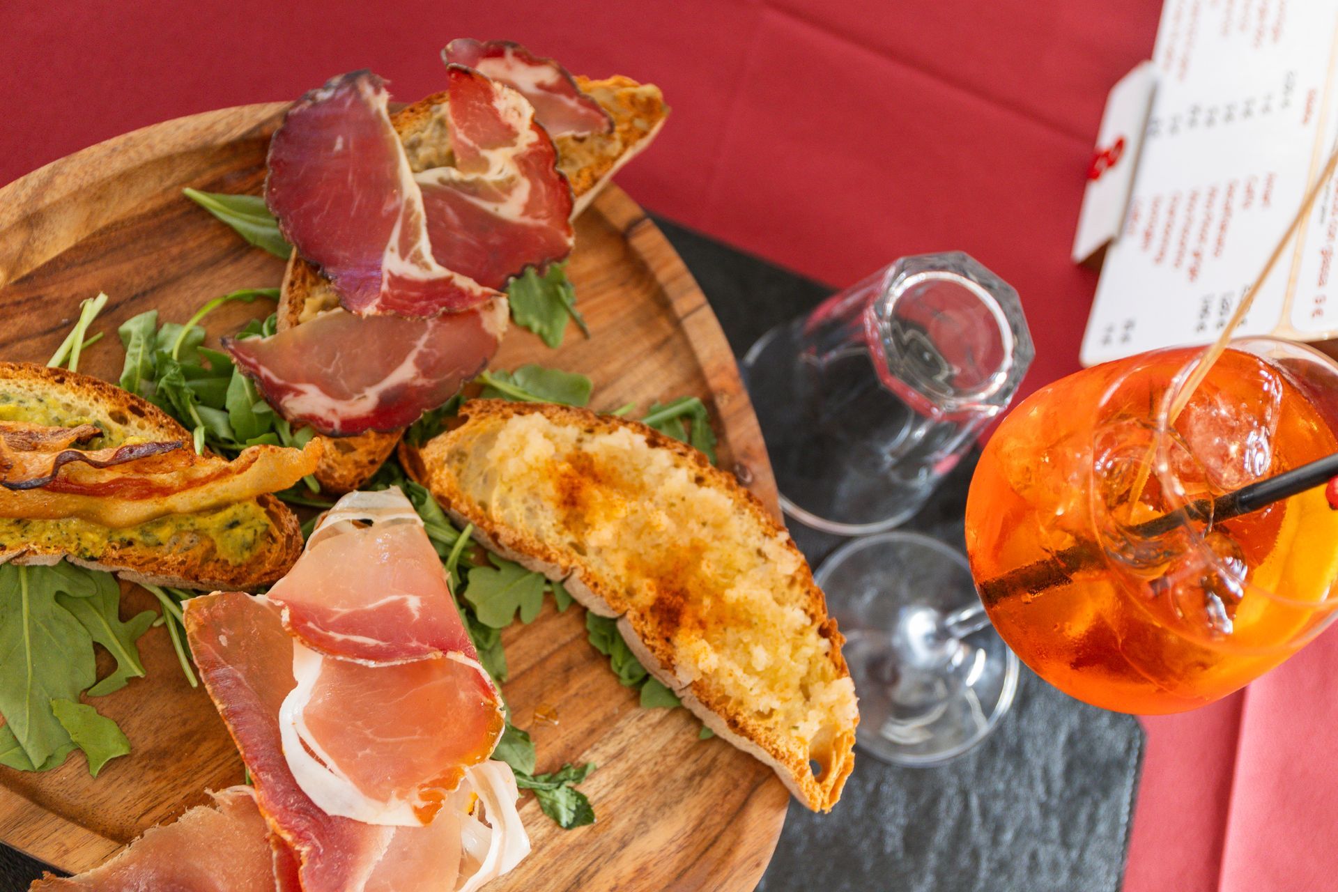 Antipasti gourmet e salumi serviti su un piatto con una bevanda arancione brillante, su una tovaglia rossa.