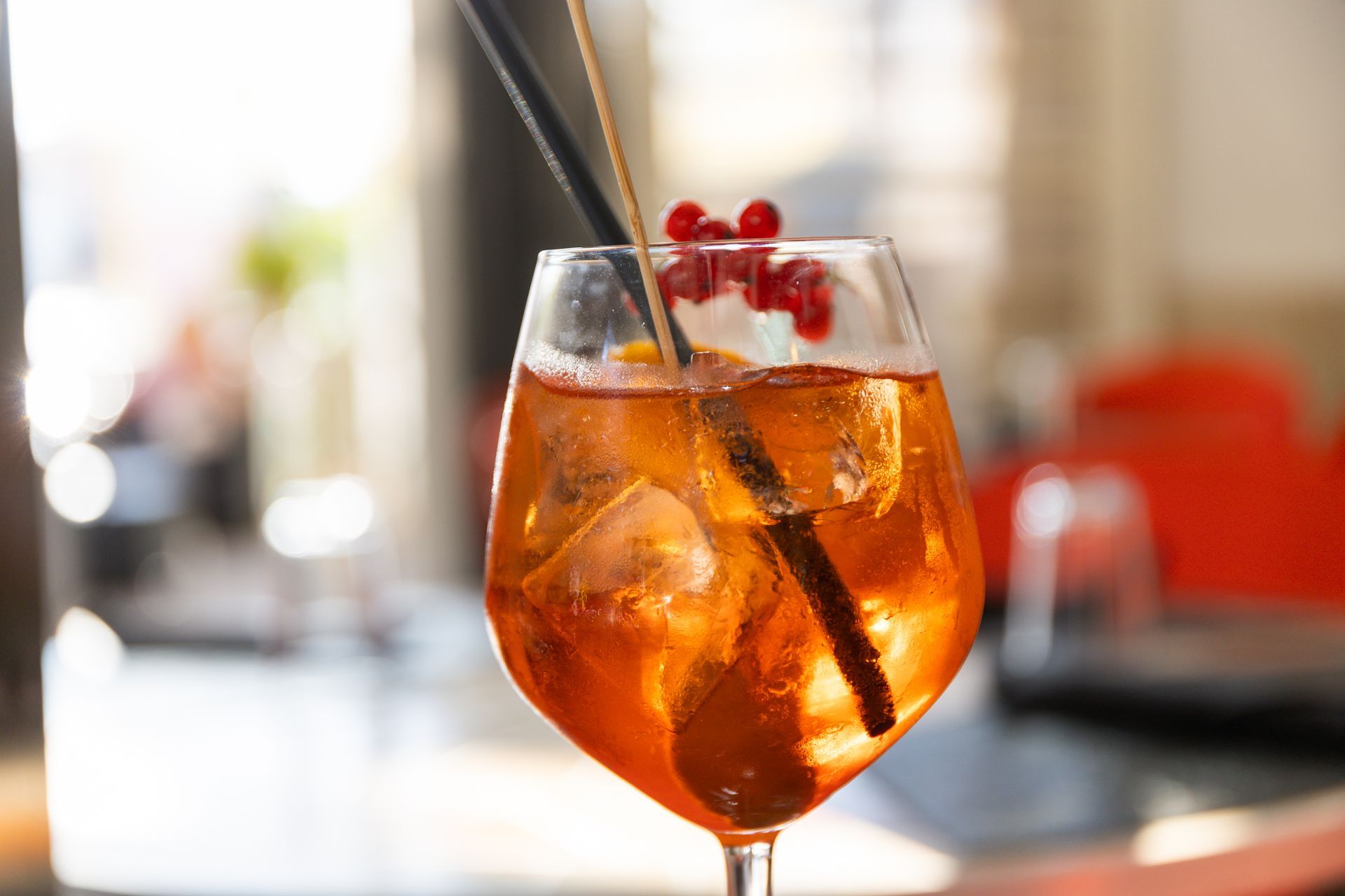 Cocktail all'arancia in un bicchiere con stelo, ghiaccio, ciliegie e cannuccia nera su un tavolo da bar.