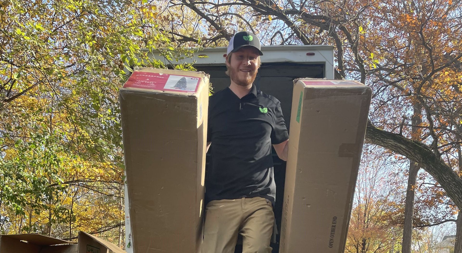 Man in black shirt and cap carries two large cardboard boxes from a truck outdoors.