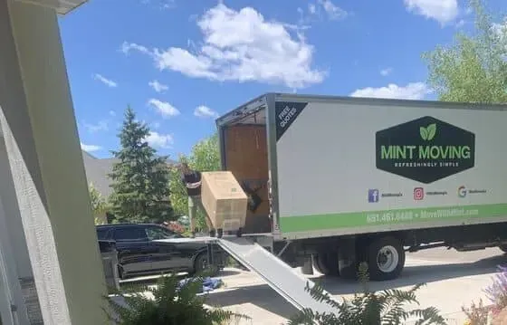 Person loading a box into a Mint Moving truck on a sunny day. A car is parked nearby.