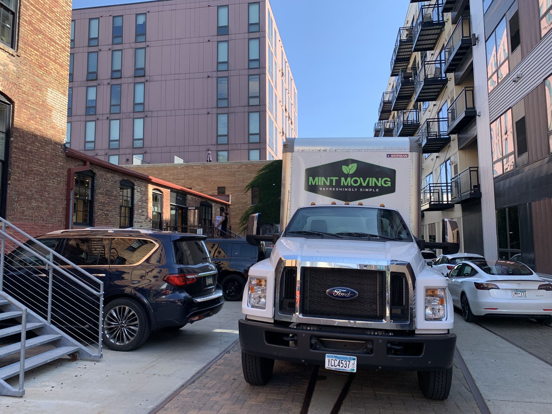 White Mint Moving truck parked in an alley; a black SUV and a white Tesla are also present.
