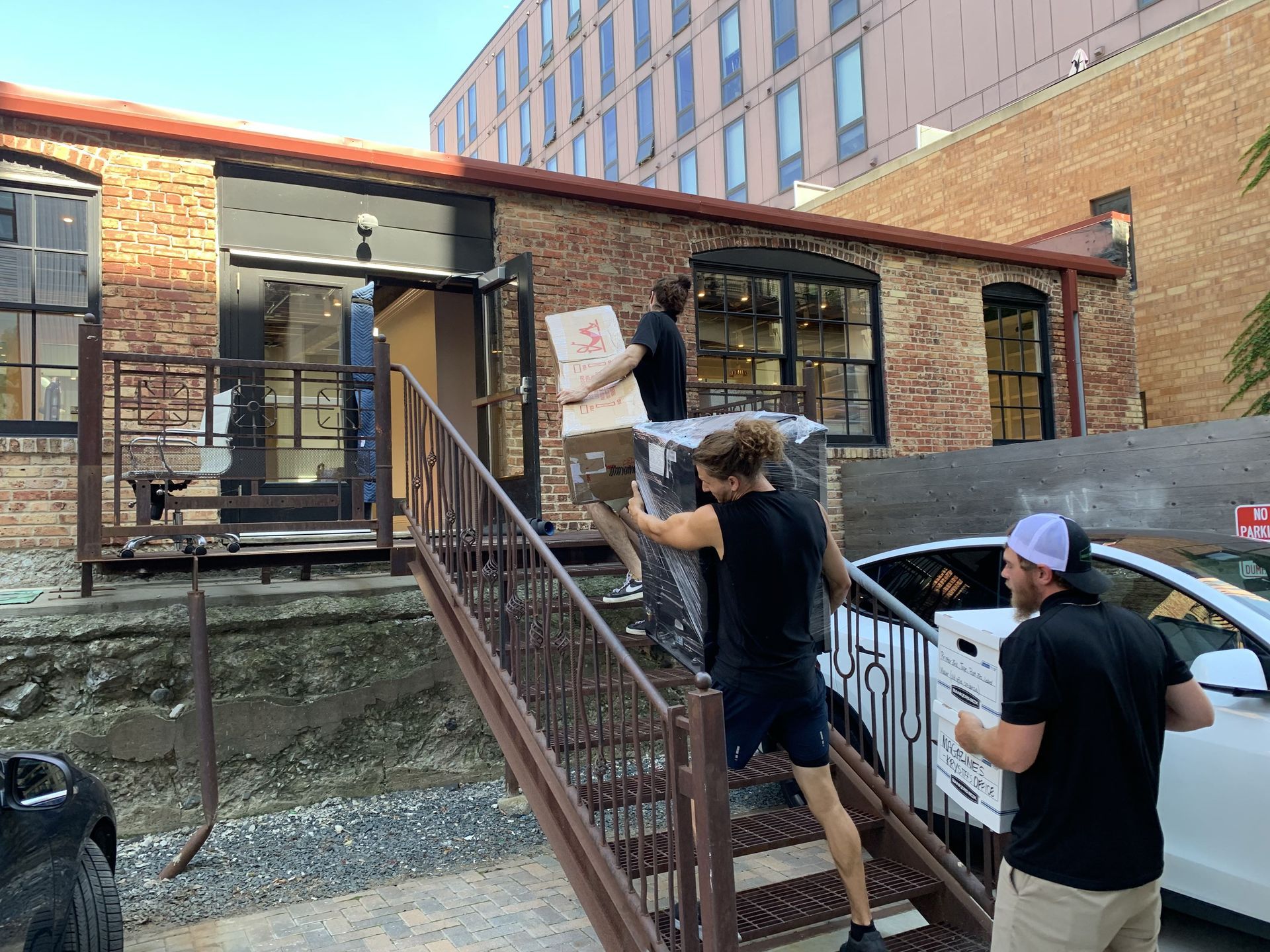 Movers carrying items up outdoor stairs into a brick building with a car parked nearby.