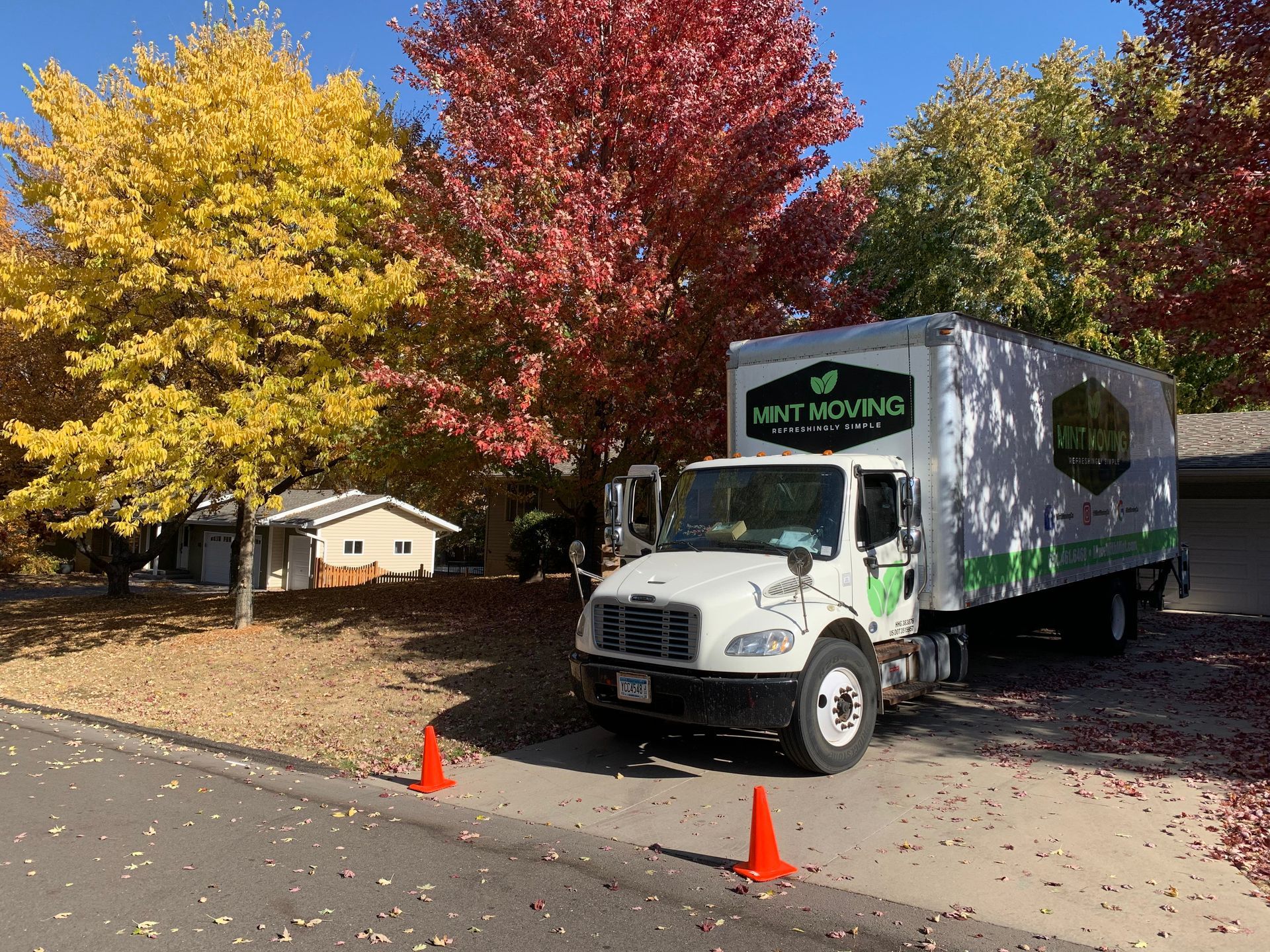 Truck parked in front of a house under trees with red and yellow leaves. Two orange traffic cones are on the ground.