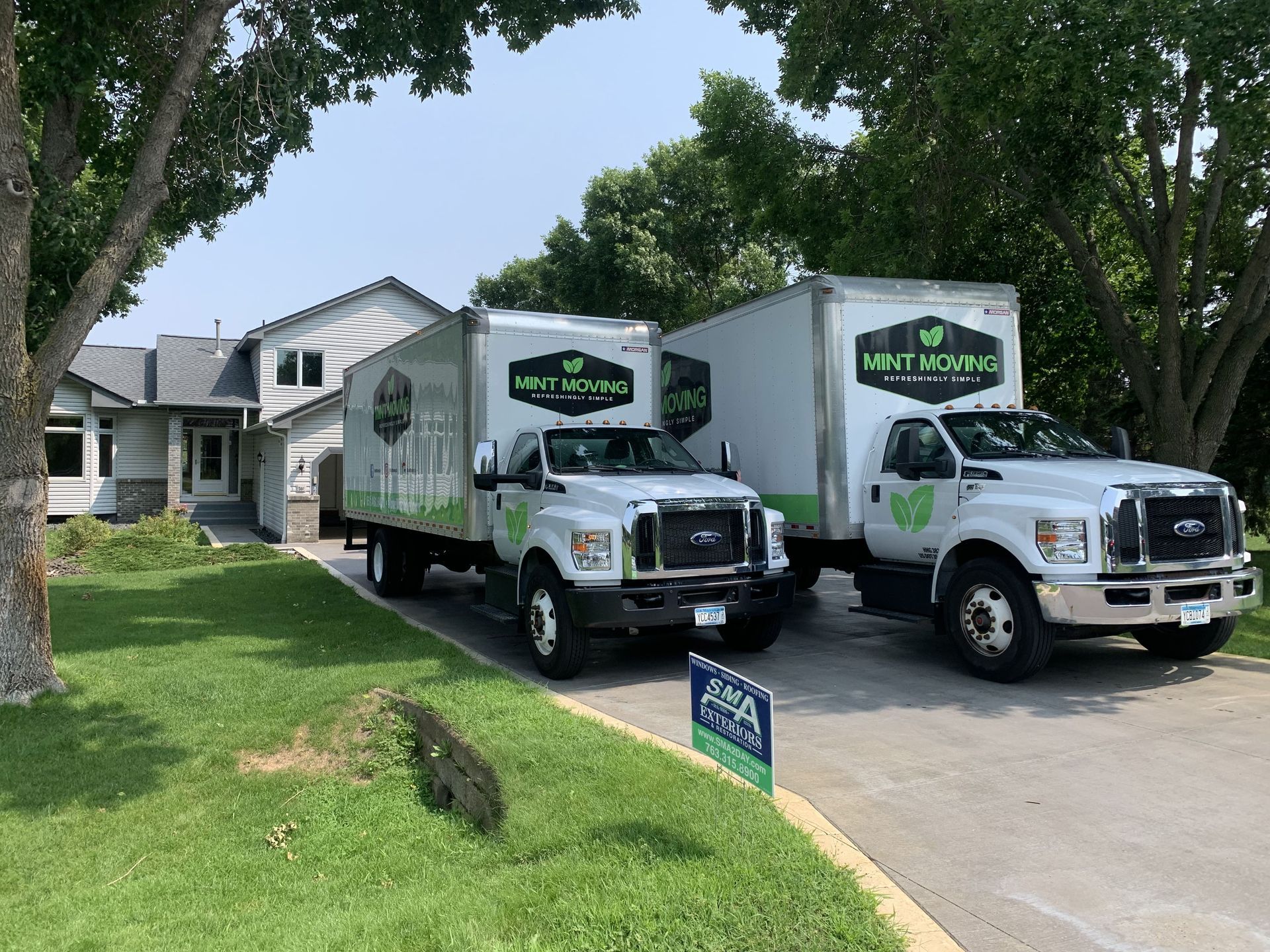 Two white moving trucks parked in front of a house, green logos on the sides, sunny day.