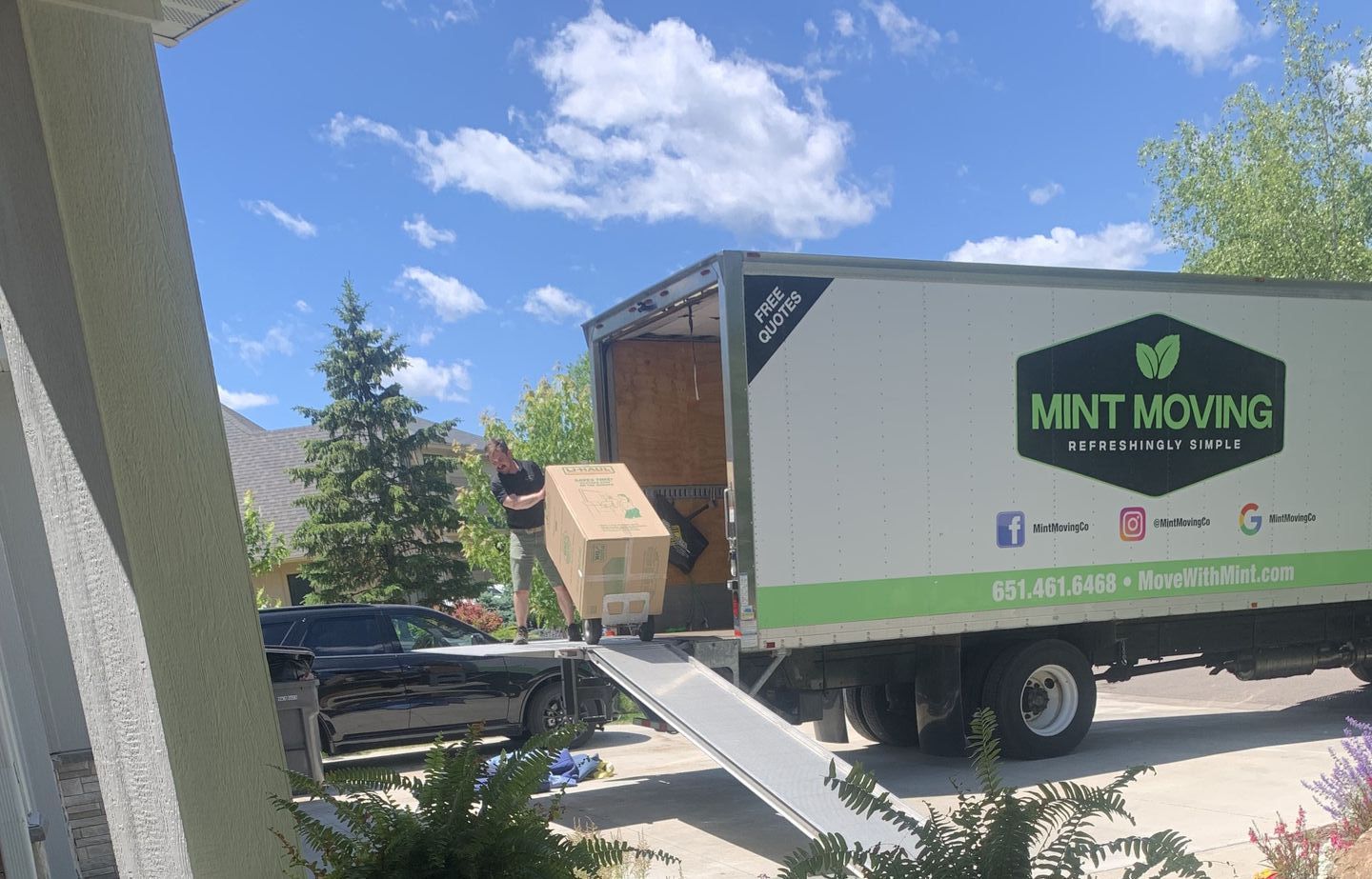A person loads a large box into a moving truck with a ramp. The truck is white with green logo. A black car is parked nearby.