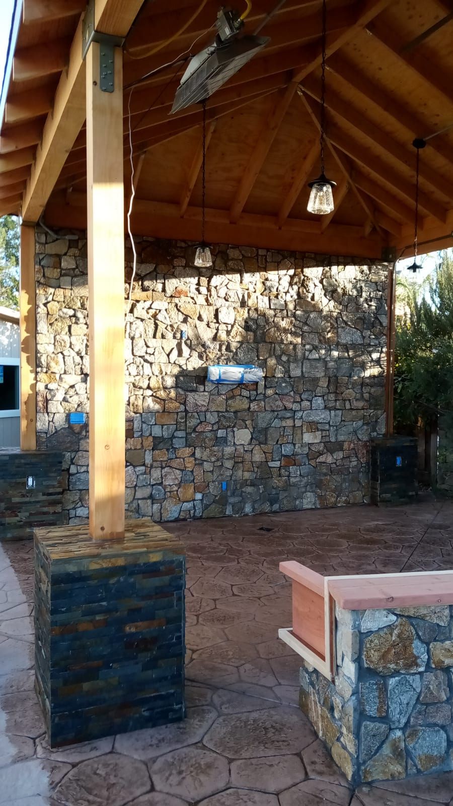 Wooden pavilion with stone walls, brick floor, and benches under a peaked roof.