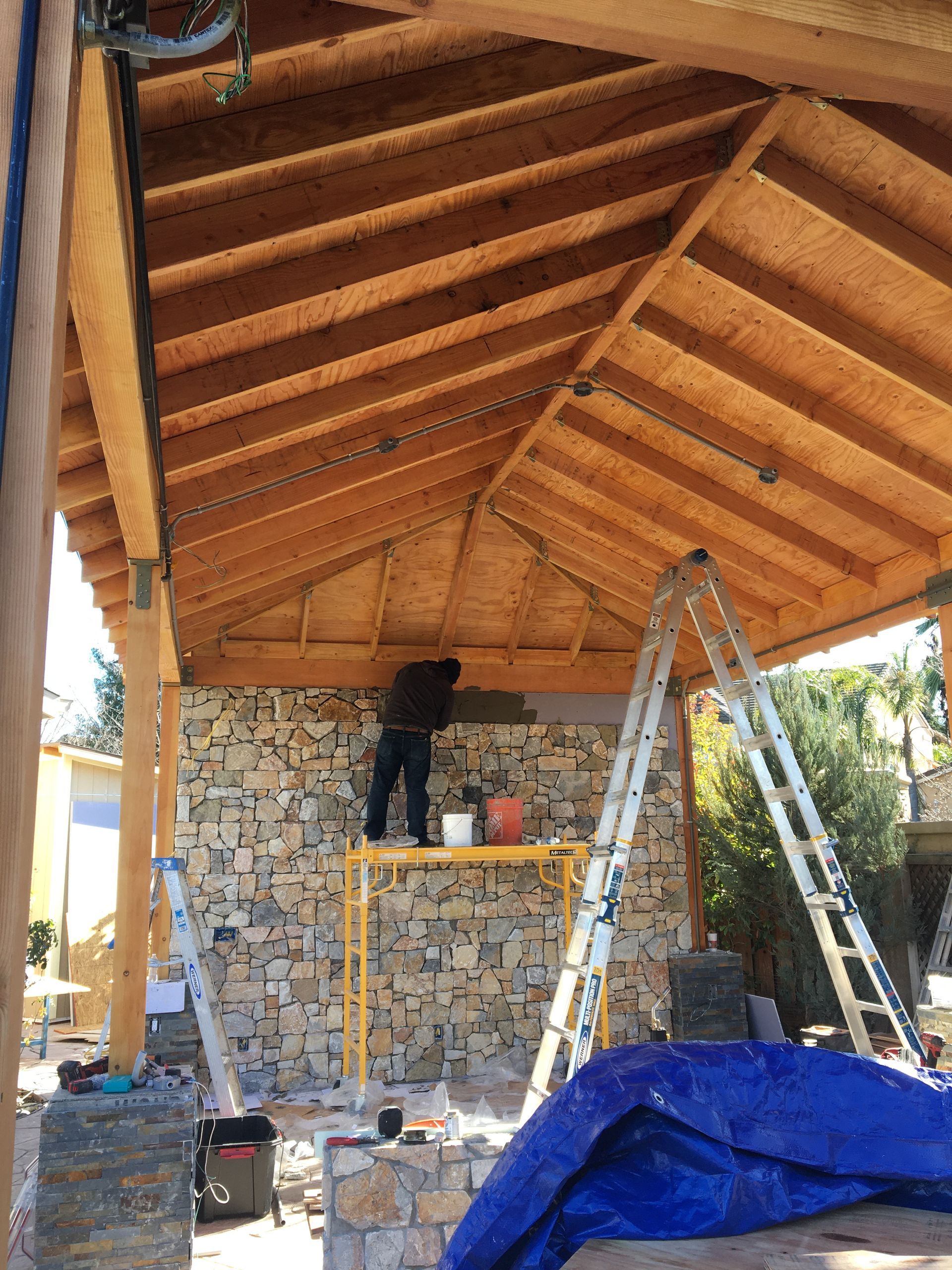 Partially built covered patio with exposed wooden rafters, stone wall, ladder, and construction materials