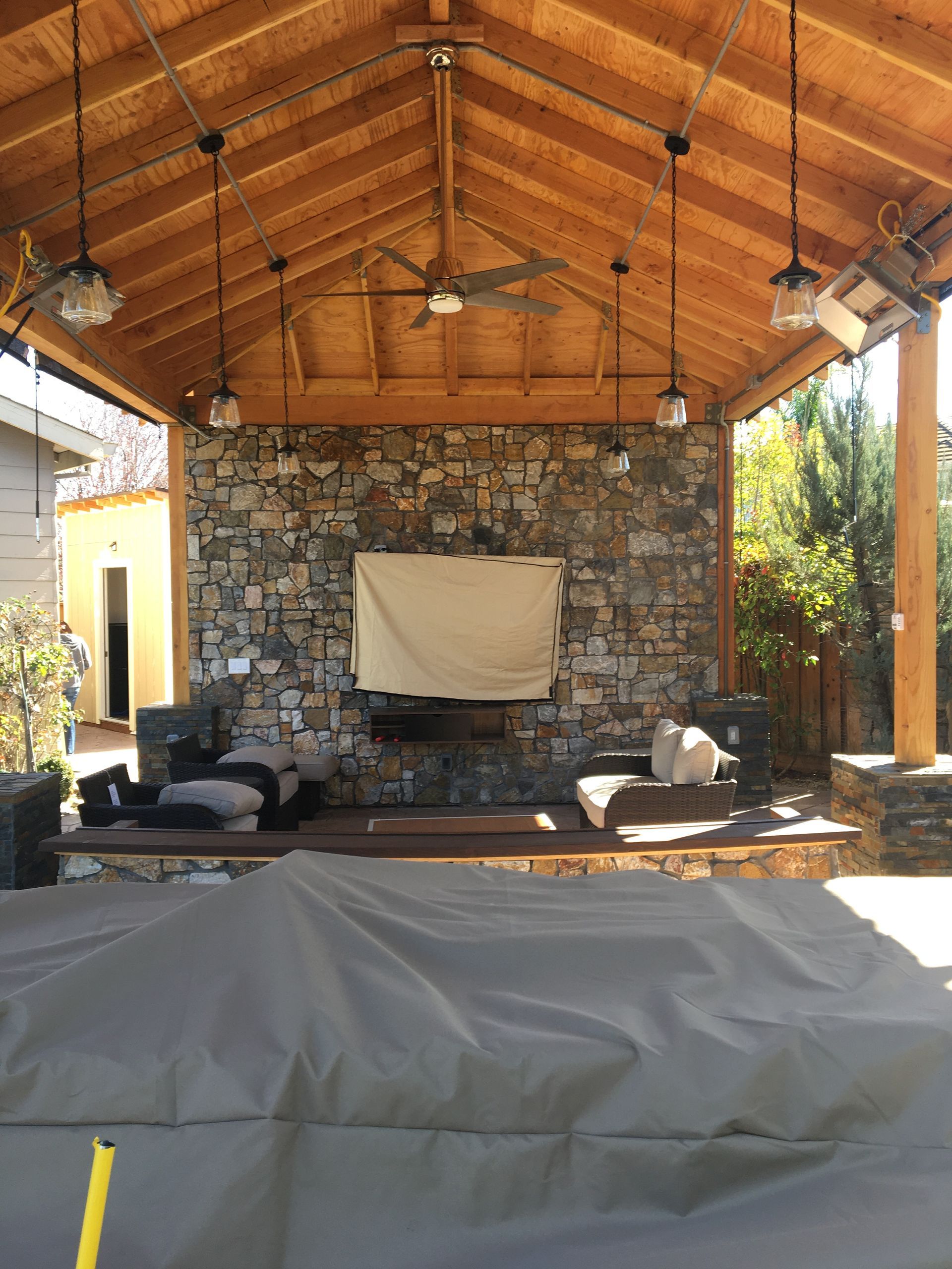 Outdoor stone fireplace under a wooden pavilion with lounge chairs and a covered table in front