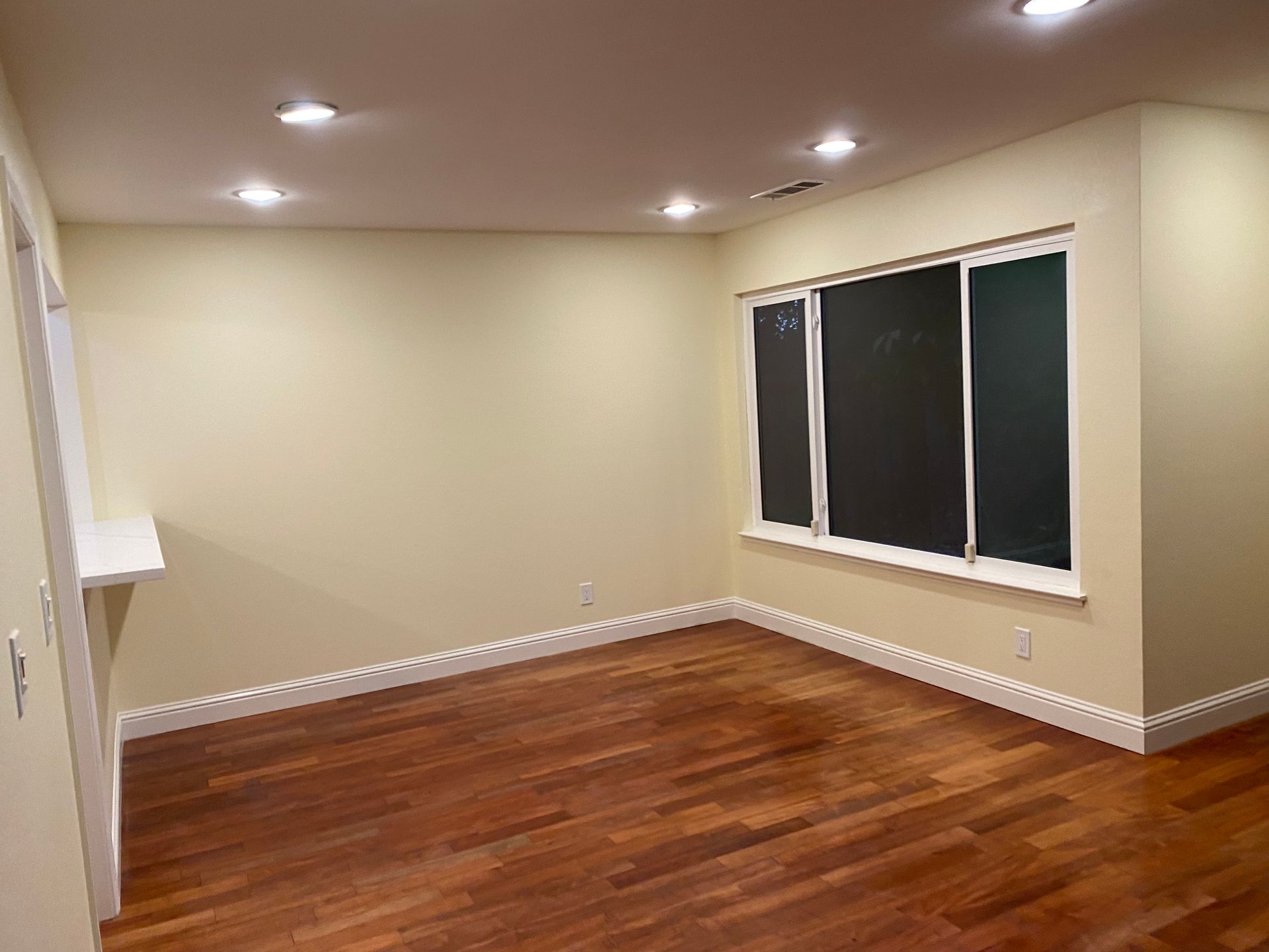 Empty beige room with hardwood floors, recessed lights, and a window with blinds