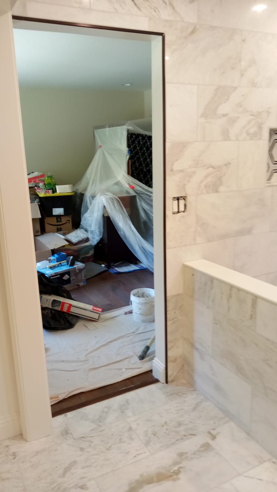 Bathroom doorway opening to a cluttered bedroom, with marble tile walls and a large mirror.