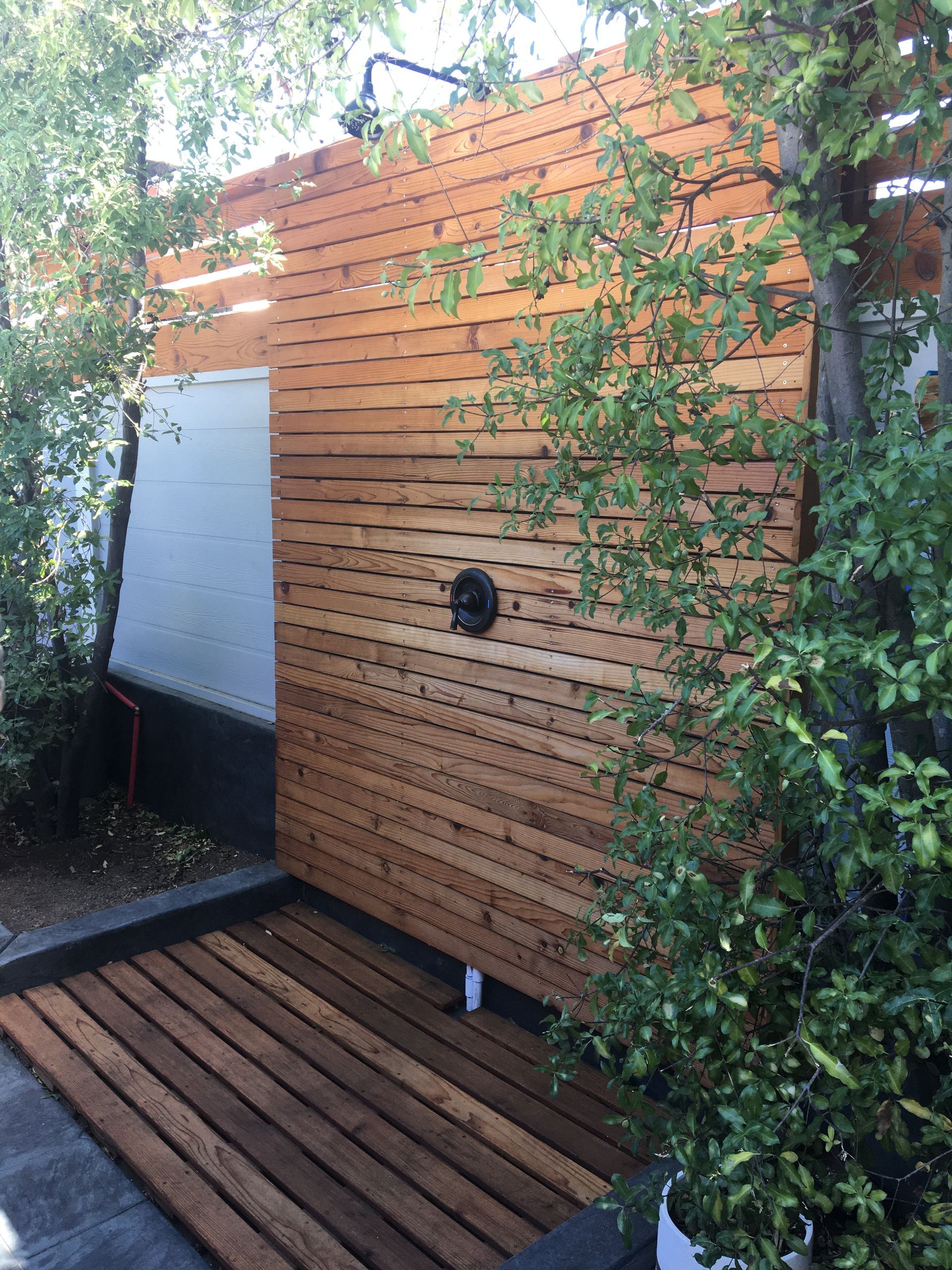 Wooden outdoor wall with horizontal slats, a white panel, and a dark deck in a leafy garden setting