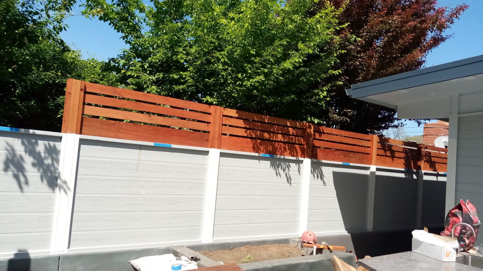 White fence with orange slatted top in a backyard beside a house and leafy trees.