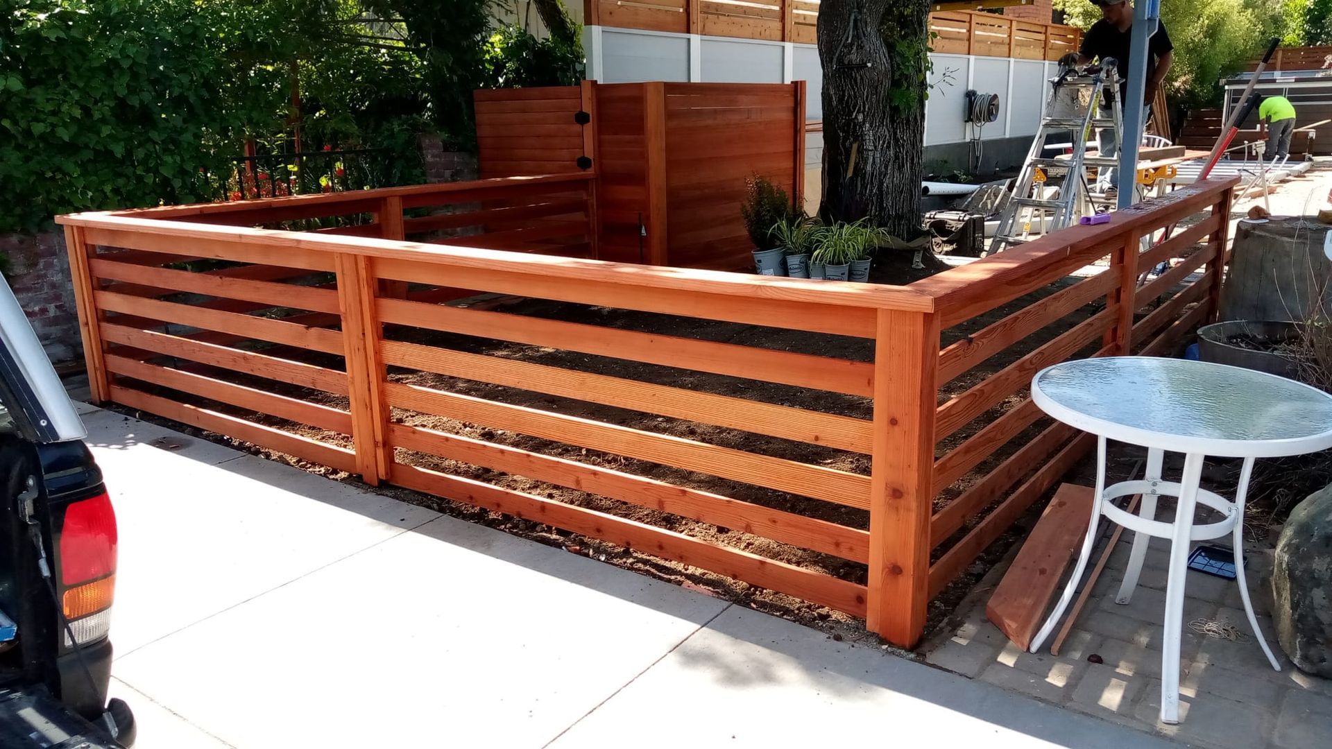 Wooden fenced patio area beside a sidewalk, with a small round metal table and parked car nearby.