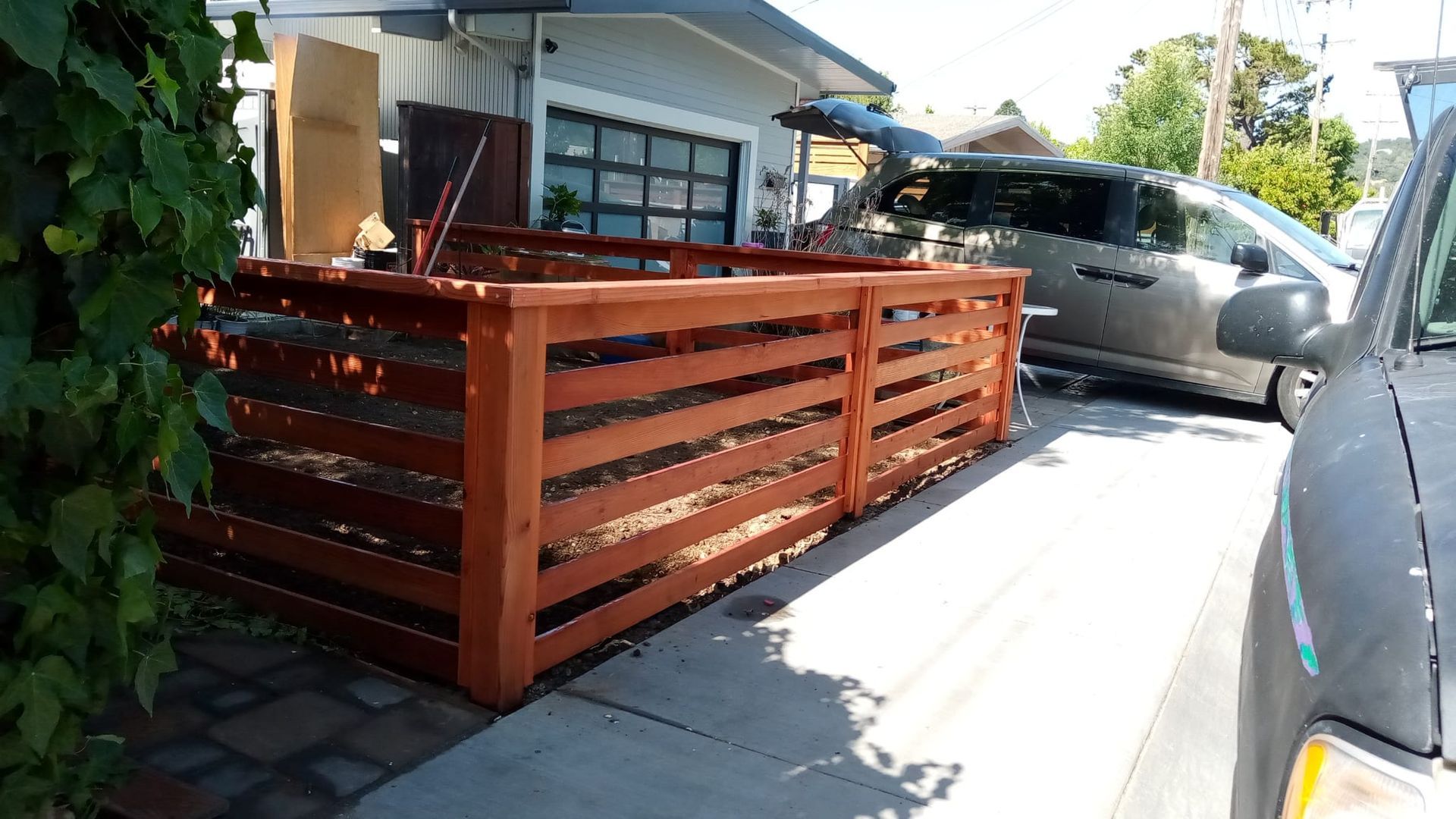 Wooden deck railing beside a driveway with parked cars and a house in the background