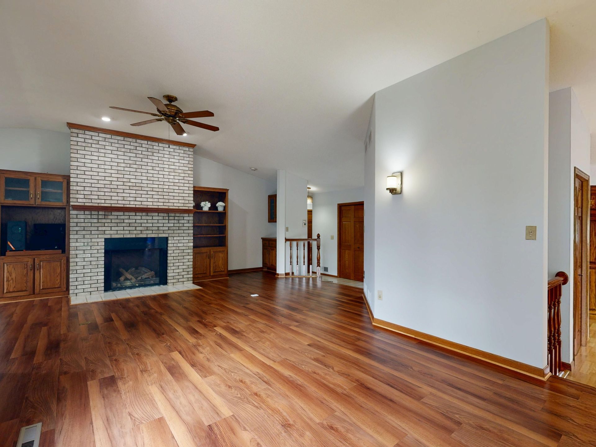 Empty living room with wood floors, brick fireplace, ceiling fan, and a white accent wall.