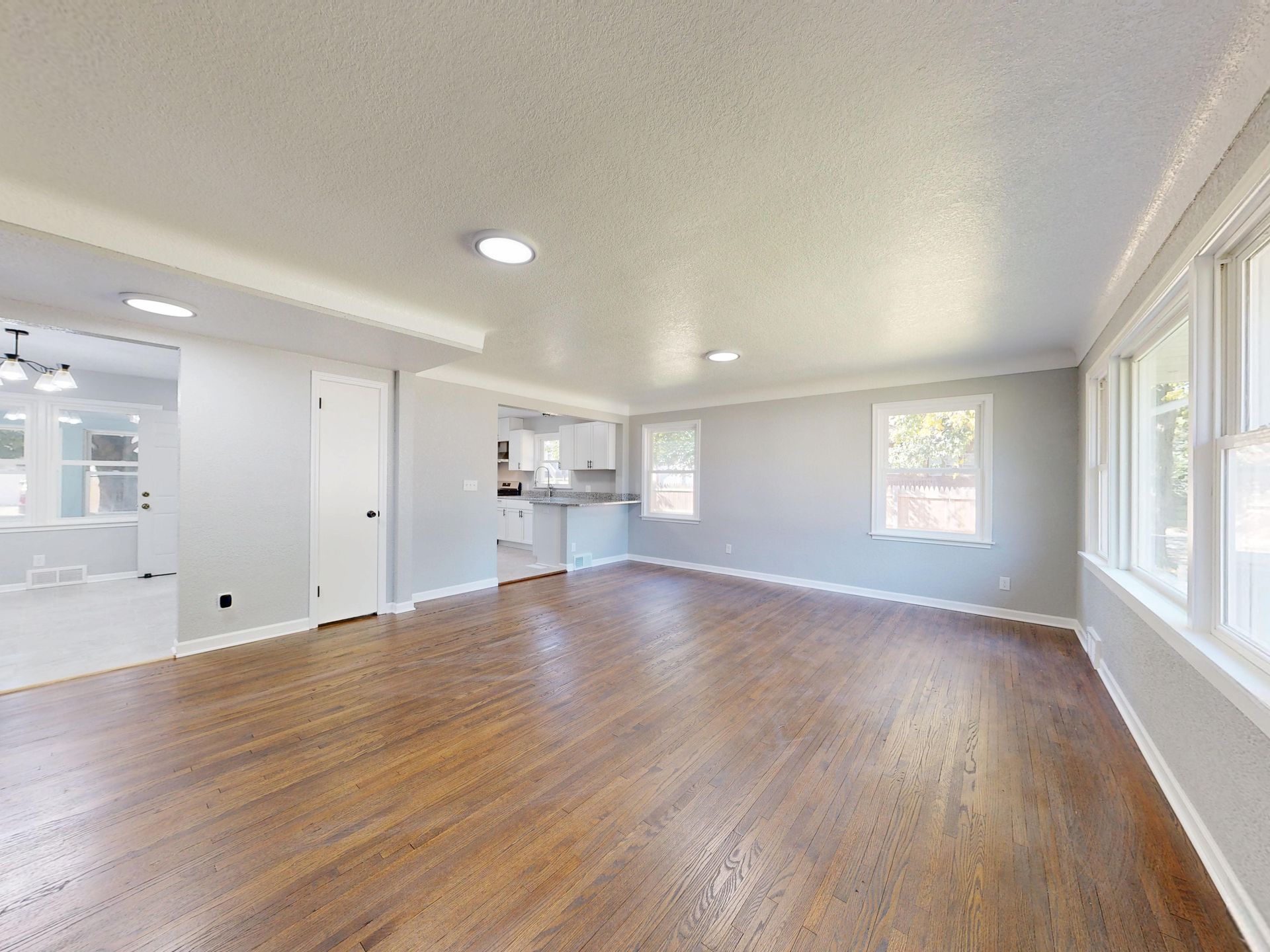 Empty living room with hardwood floors, white walls, recessed lights, and large windows.